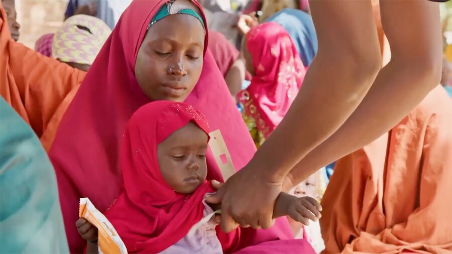 A woman in a pink headscarf holds her daughter as a health worker wraps a plastic band around the little girl's wrist to test for malnutrition. Photo: WFP/Arete/Nommiyid Chantu