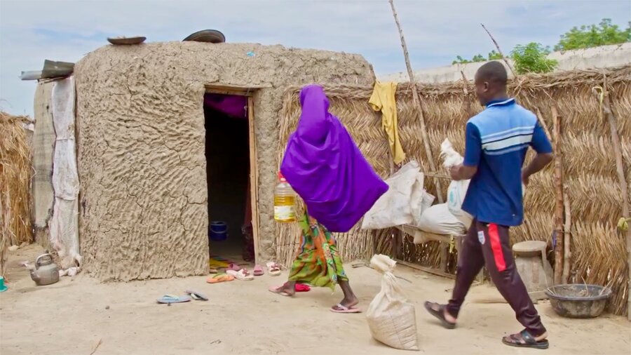 A woman in a purple headscarf and a man in shorts and a blue top carry bags of WFP food into a mud-brick hut at a displacement camp in northeastern Nigeria. Photo: WFP/Arete
