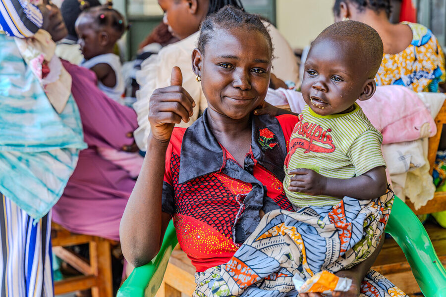 A woman looking sad and relieved holding a toddler as she gives a thumbs up at a distribution point in DRC