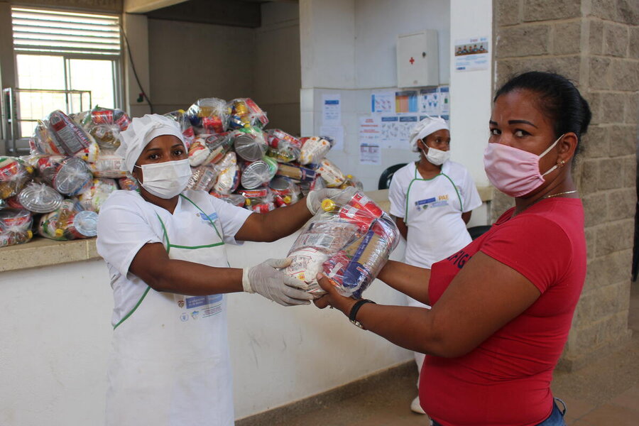 A woman in kitchen overalls hands a pack of food to a woman in re t-shirt, both wear covid face masks