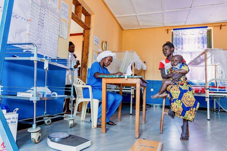 A woman wearing a red T-shirt and colourful skirt brings her baby daughter into a hospital for a checkup. Photo: WFP/Charly Kasareka