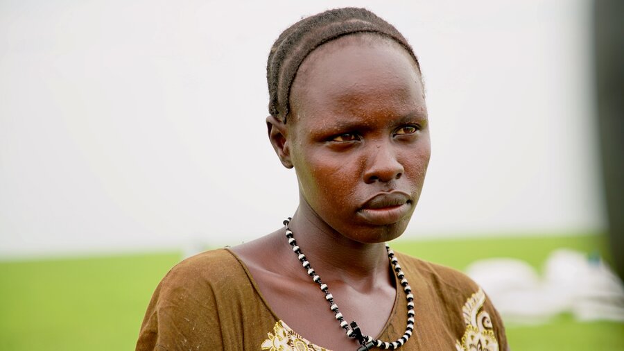 Closeup of a woman's face, which looks sad, backdropped by a green field and white food bags. Photo: WFP/Eulalia Berlanga