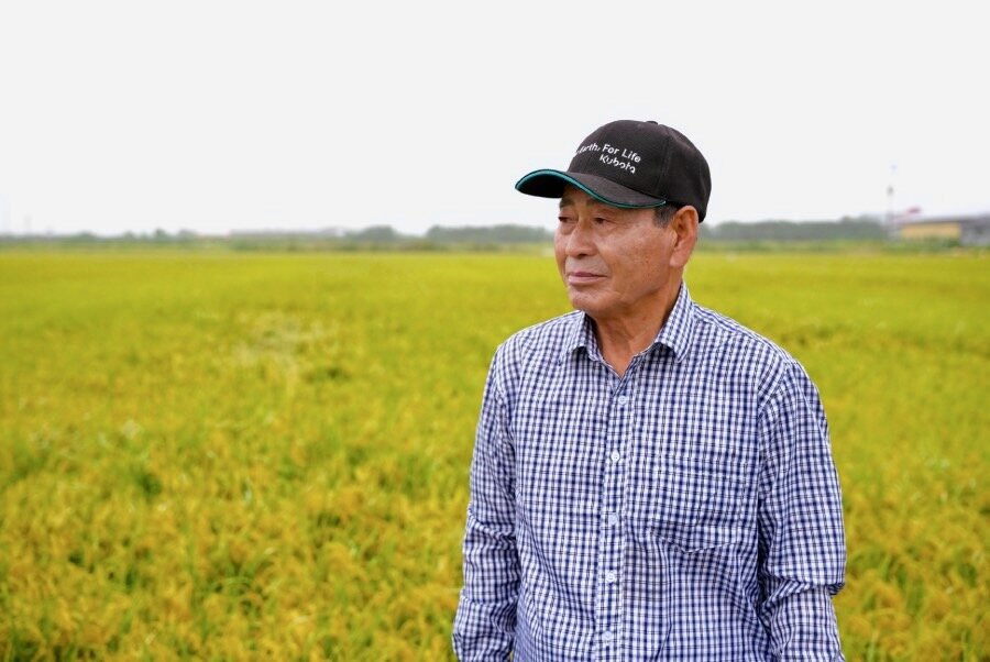 An elderly farmer sporting a baseball cap and blue checked shirt stands in front of his flowering rice field. Photo: WFP/Yanghae Won