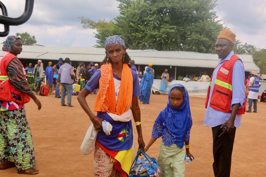 A woman with an orange shawl and sad expression holds the hand of a little girl as she walks past staff at a refugee camp. Photo: WFP
