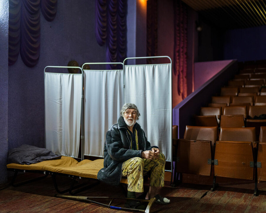 Bearded man in green jacket sits on a bed in front of medical screen curtains, tiered auditorium seats visible on his right