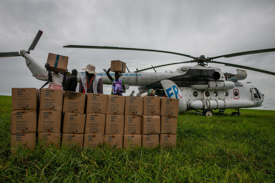 A white helicopter marked WFP is parked on green grass under a cloudy sky. Several people unload cardboard boxes labeled “Relief Items,” stacking them in front of the aircraft for distribution.