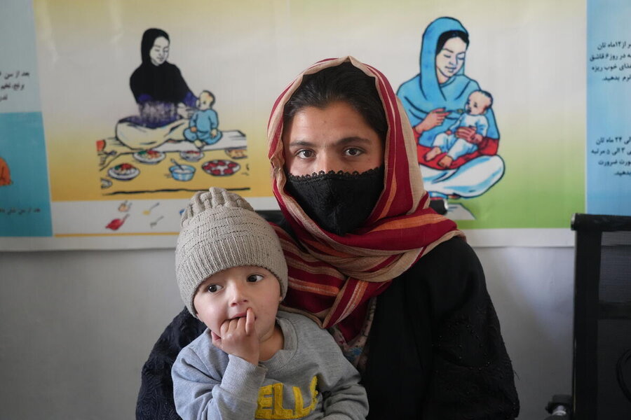 A woman with a face covering in Afghanistan holds a child with informercial illustrations pined to the wall behind her