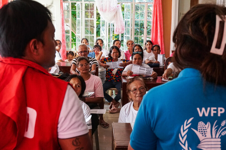 Two humanitarians with their backs to the camera, one in a WFP shirt, address a small room of people