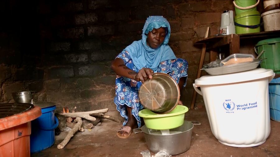 A young woman in a light blue veil sits on an earthen floor, pouring ingredients for a nutritious porridge into a bowl. Photo: WFP