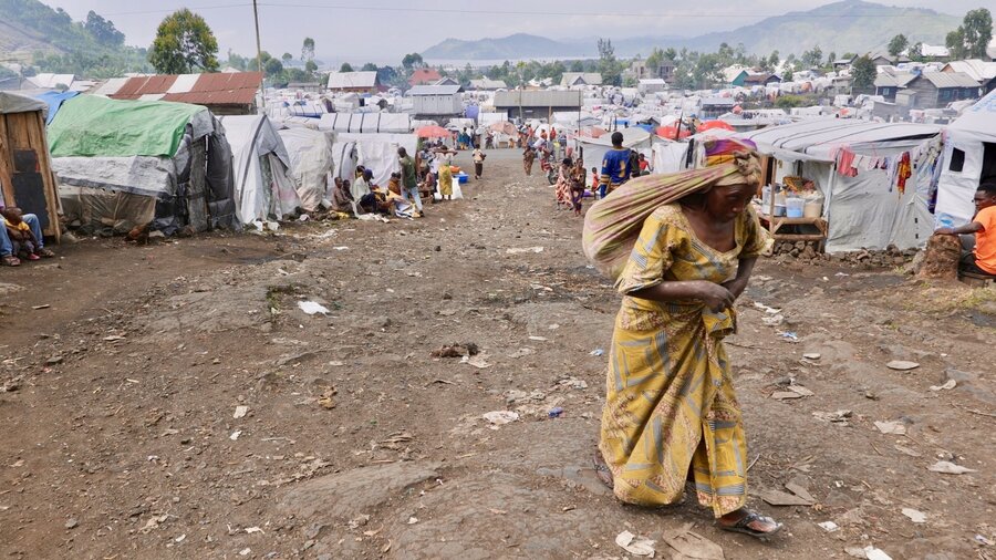 A woman in a yellow dress, carries a bundle strapped on her forehead, as she slogs up a dirt path - backdropped by a makeshift displacement camp. 