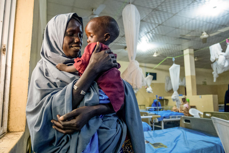 A woman in a long headscarf lovingly holds her little son, wearing a red hospital gown and blue diapers. Photo: WFP/Nommiyid Chantu