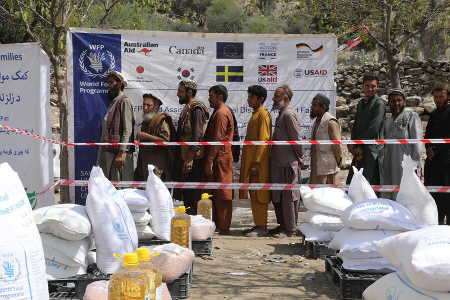 Men in a line separated by red marker tape from food packs in the forground