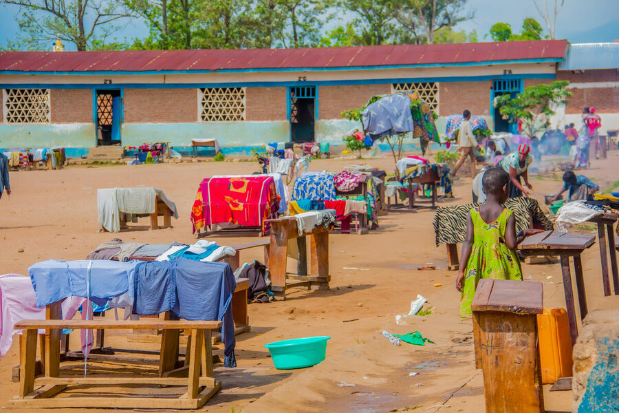 A little girl and woman at a makeshift outdoor shelter in eastern DRC - a collection of furniture and belongings outside a low building. Photo: WFP/Biobei Malenga 