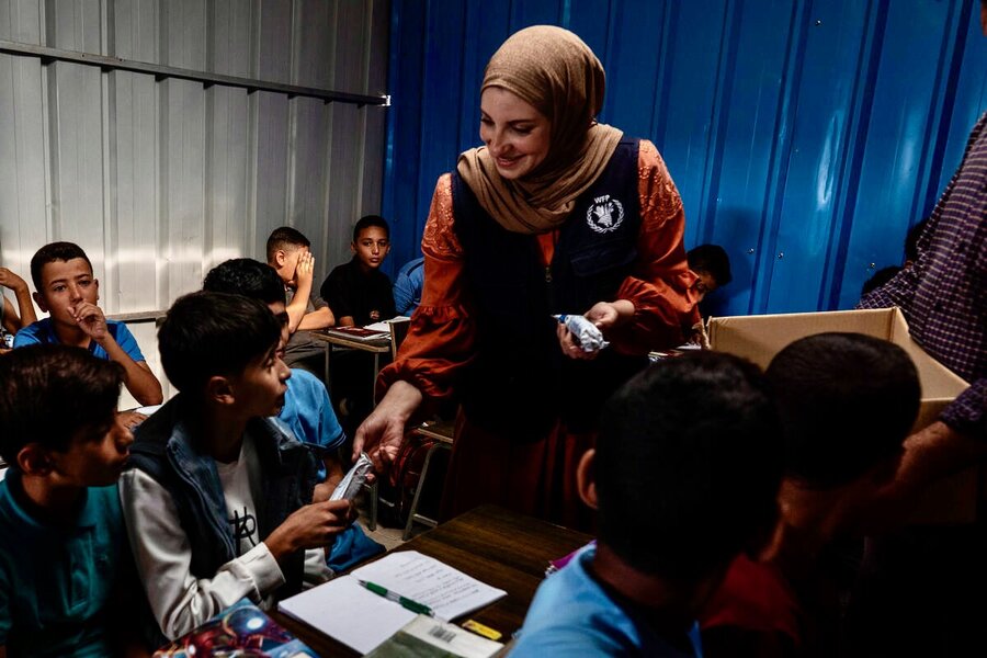A smiling WFP staffer in a blue vest and tan headscarf hand out snacks to young students in a shipping container turned makeshift classroom in Deir al-Balah, Gaza. Photo: WFP/Nour Hamad