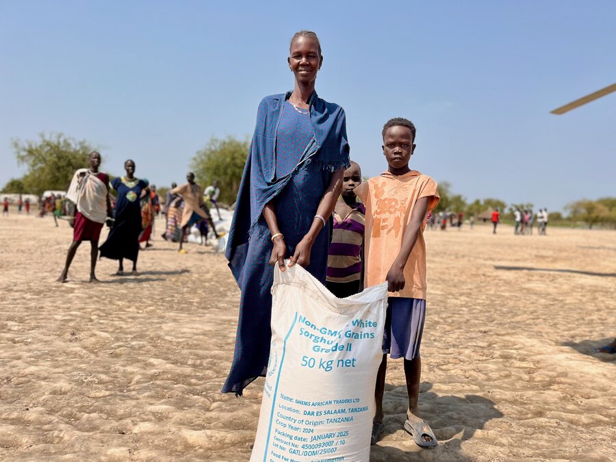 A smiling mother and child holding a bag of WFP food, as they stand in a sandy stretch of land backdropped by trees and people. Photo: WFP/Hedley Tah