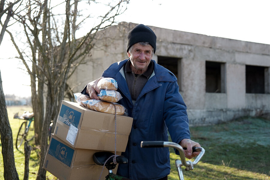 Man pushes a bicycle loaded with WFP food boxes and bread packages while standing near an abandoned building in a rural area.