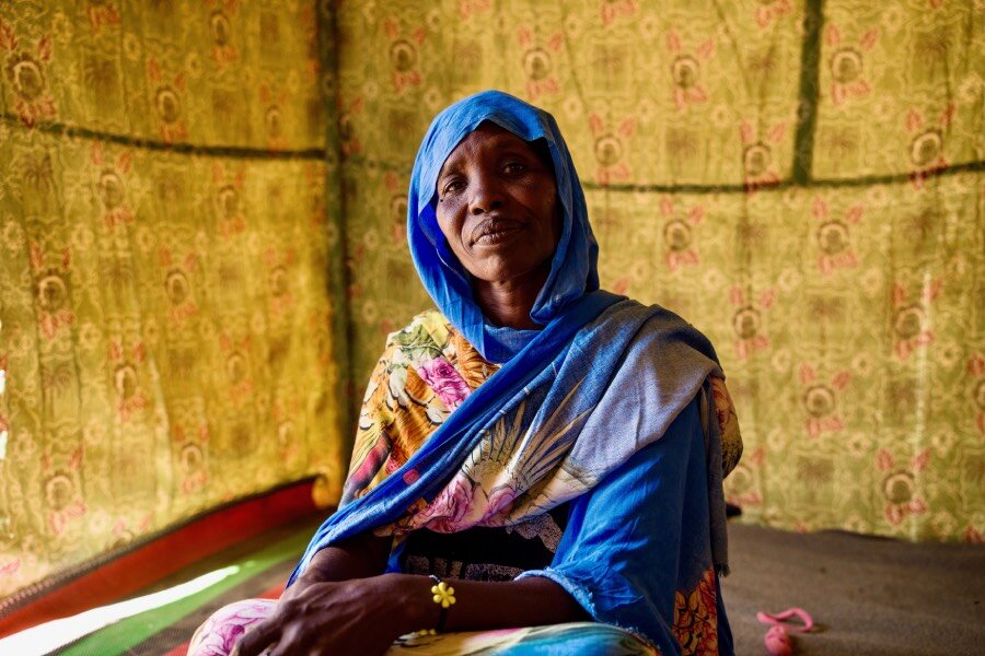 A woman in a blue patterned headscarf sits in a makeshift cloth tent. Photo: WFP/Abubakar Garelnabei