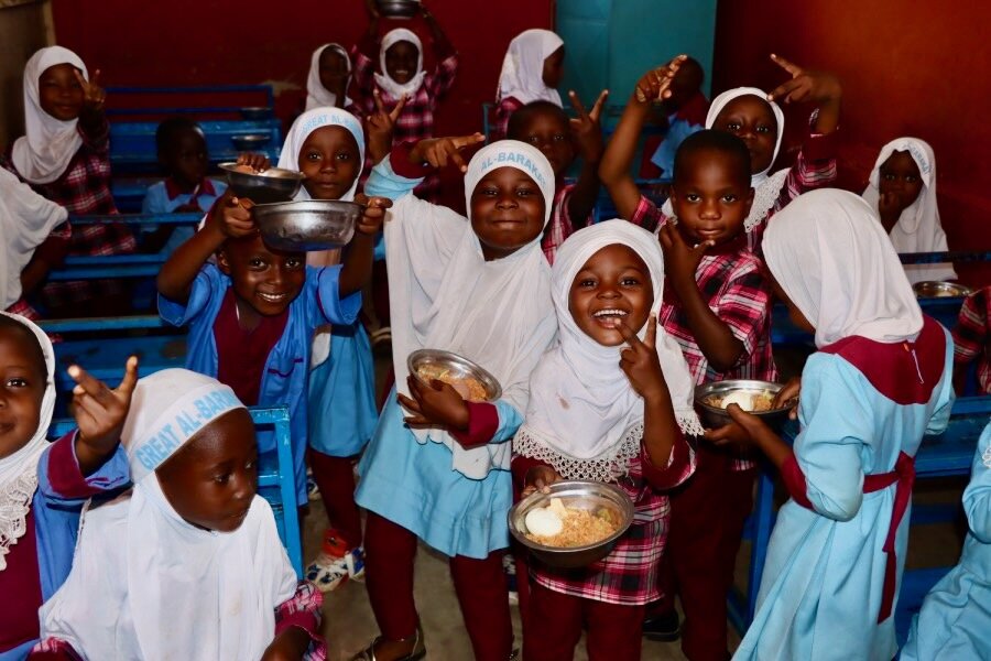 Smiling little girls in blue uniforms and white headscarves thrust up metal bowls of food. Photo: WFP/Abdul-Wahab Mohammed