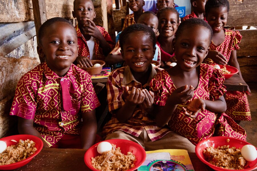 Smiling girls and boys in red patterned uniforms with plastic plates of food in front of them. Photo: WFP/Abdul-Wahab Mohammed 
