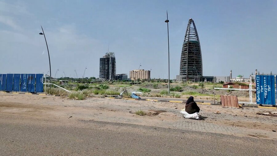 A woman sits next to a dusty road, backdropped by war shattered buildings. Photo: WFP/Philippe Kropf
