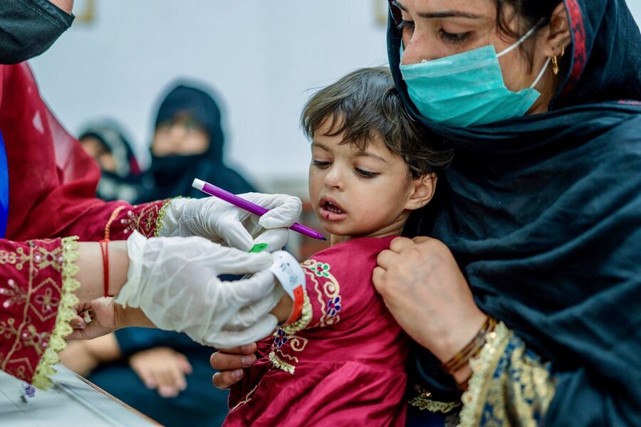 A mother in a robe and face mask holds a little girl who is getting her mid-arm checked for malnutrition by a health worker via a special band. Photo: WFP/Sahil Zia