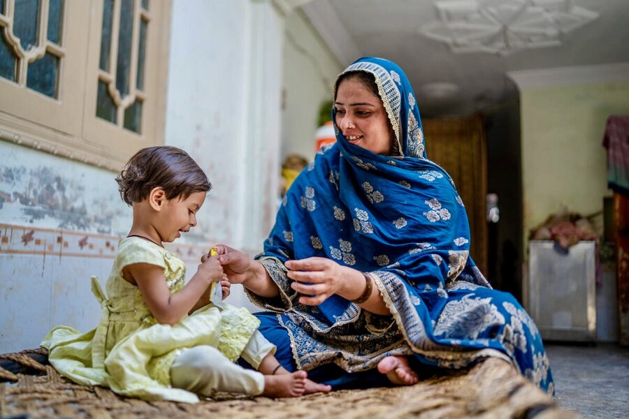 A mother in a long blue headscarf and robe hands a smiling little girl in a yellow dress a package of specialized nutritional food. Photo: WFP/Sahil Zia