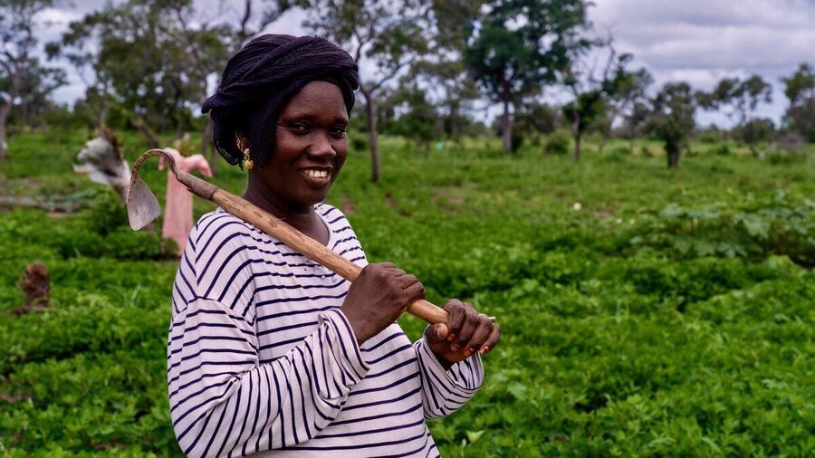 A smiling woman stands in a lush green field holding a hoe. Photo: WFP/Sylvie Njobati