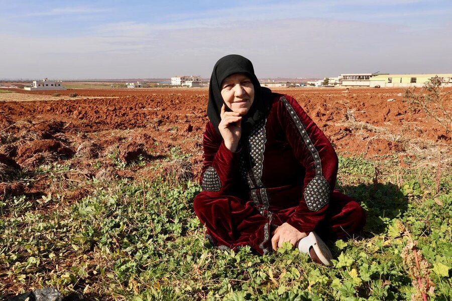 A woman in a dark red gown and black headscarf sits in the middle of a field backdropped by a housing development. Photo: WFP/Zuha Akkash