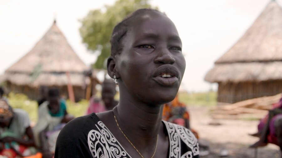 A head shot of a woman in a black patterned top, backdropped by straw and reed huts and people. Photo: WFP/Gabriela Vivacqua