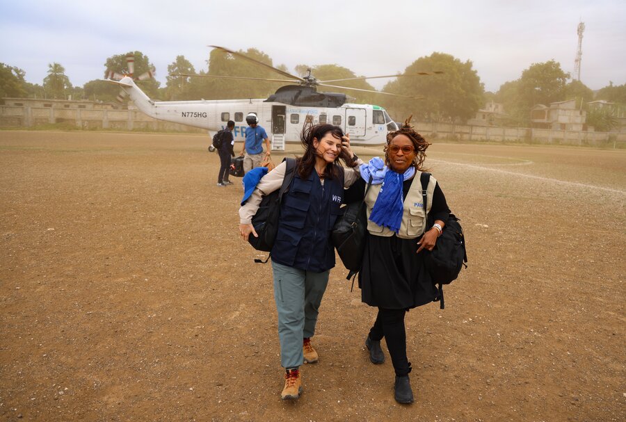Two women with WFP vests walk down a dusty field with a WFP helicopter in the background. Photo: WFP/Emily Pinna