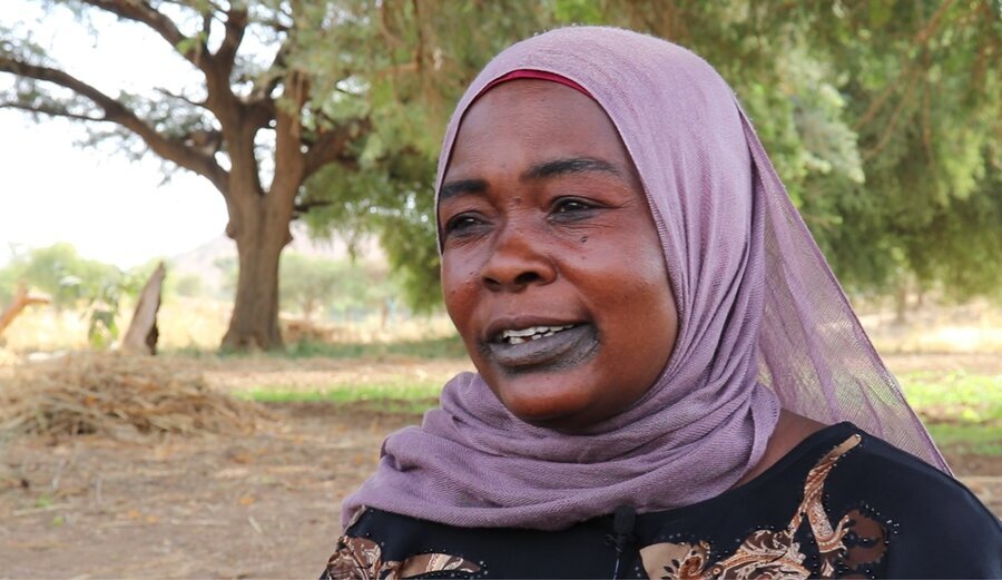 A closeup of a smiling woman in a purple headscarf standing in a sandy field with trees in the background. Photo: WFP/Petroc Wilton