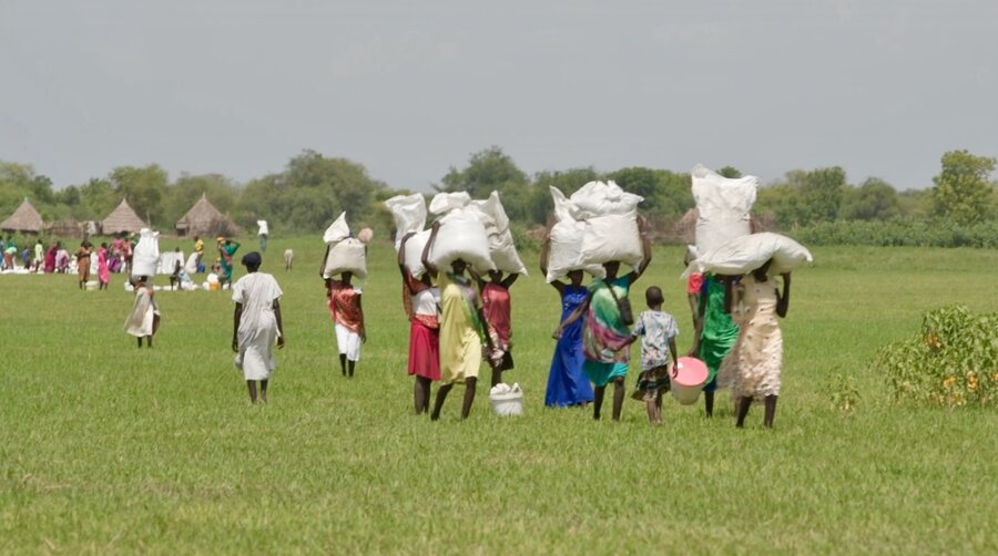 People walking through a green field balancing large white bags on their heads. Photo: WFP/Gabriela Vivacqua