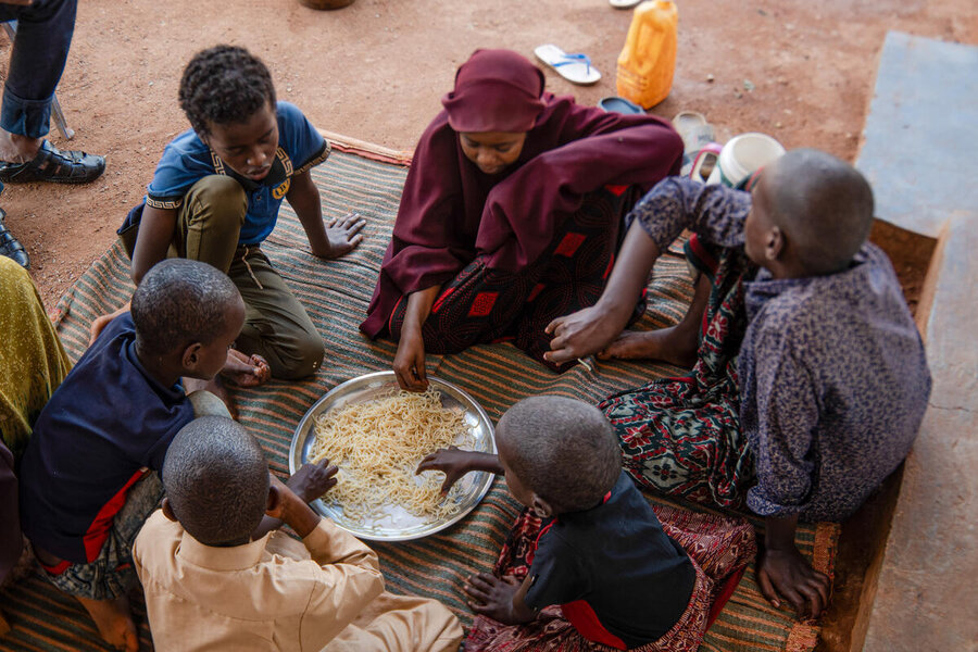 Children sit in a circle on a mat, sharing cooked noodles from a metal tray outdoors, using hands to eat together at a temporary relief site setting.