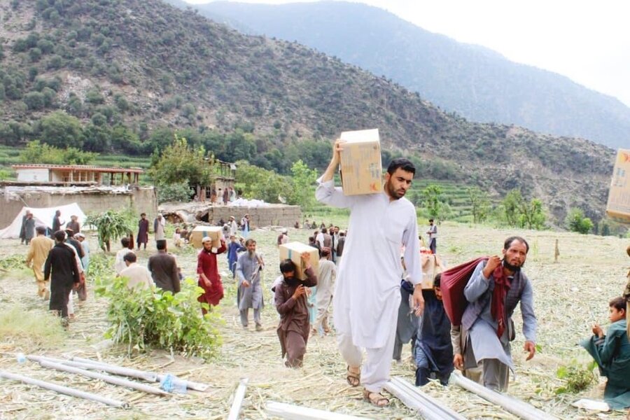 People carry cardboard boxes uphill through a rural village, crossing fields with mountains in the background during a distribution of aid supplies.