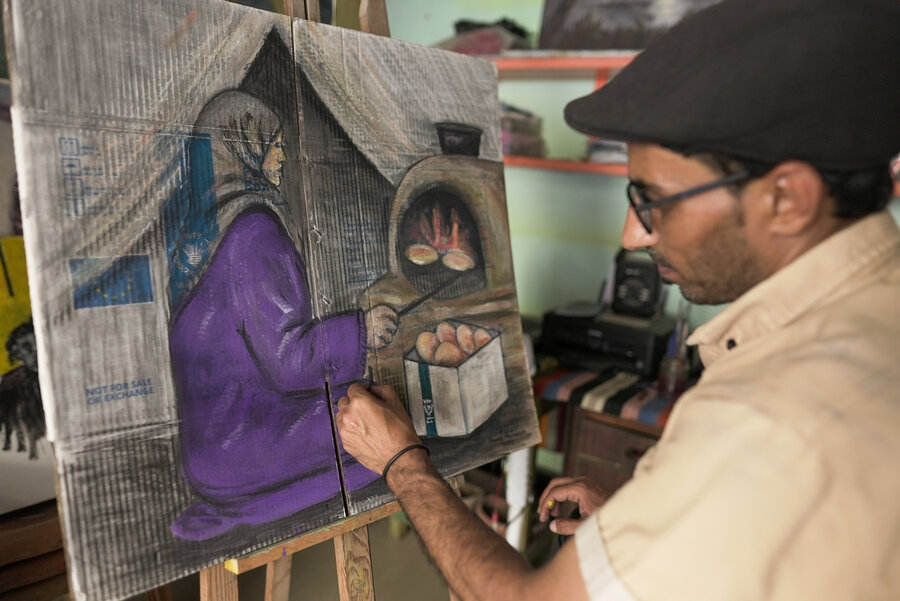 An artist wearing a beret and tan shift paints a woman in headscarf and long purple gown baking bread that's stored in a WFP food box. Photo: WFP/Jaber Badwan