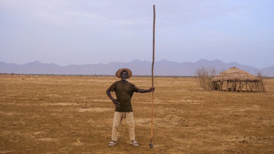 A man wielding a large stick up high stands in parched land with a hut and hills in the background. Photo: UNEP Malachie Nabbi