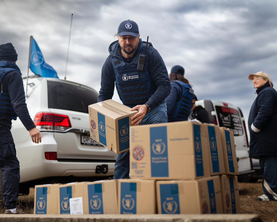 A man in a blue WFP cap and jacket unloads boxes of WFP food under cloudy skies, backdropped by people and vehicles. Photo: WFP/Sayed Asif Mahmud