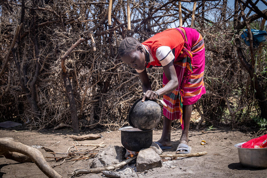 A woman in a red vest and colorful skirt cooks over an open fire, backdropped by a dry landscape. Photo: WFP/Daisy Masembe