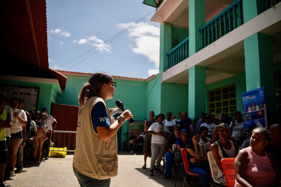 A woman with a tan WFP vest and blue T-shirt speaks to a crowd of people sitting in plastic chairs sitting in the courtyard of a green-and-white building. Photo: WFP/Gustavo Vera