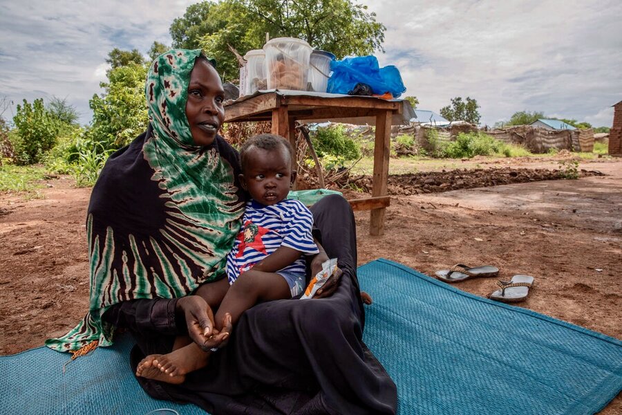 A Sudanese refugee, cradling her small son, sits outside on the ground in front of her small wooden stand, where she sells snacks to fellow refugees. Photo: WFP/Gabriela Vivacqua