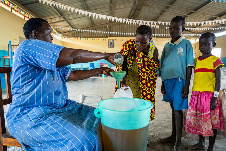 A woman wearing a blue dress pours oil into a plastic container held out by another woman, with her two children standing nearby. Photo: WFP/Daisy Masembe