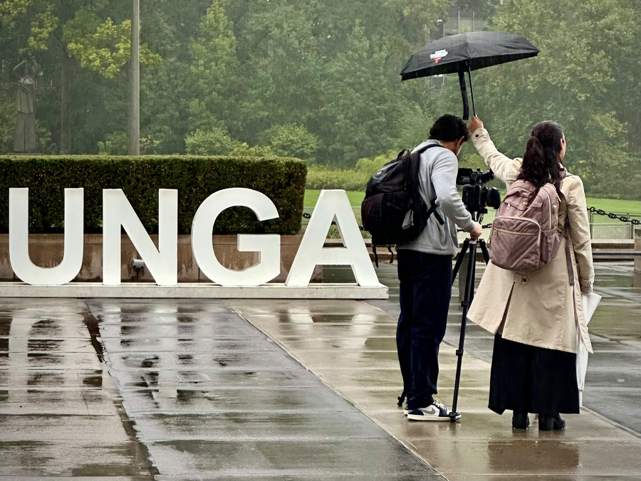 Two people stand beside a camera in the rain near large UNGA letters outside the venue