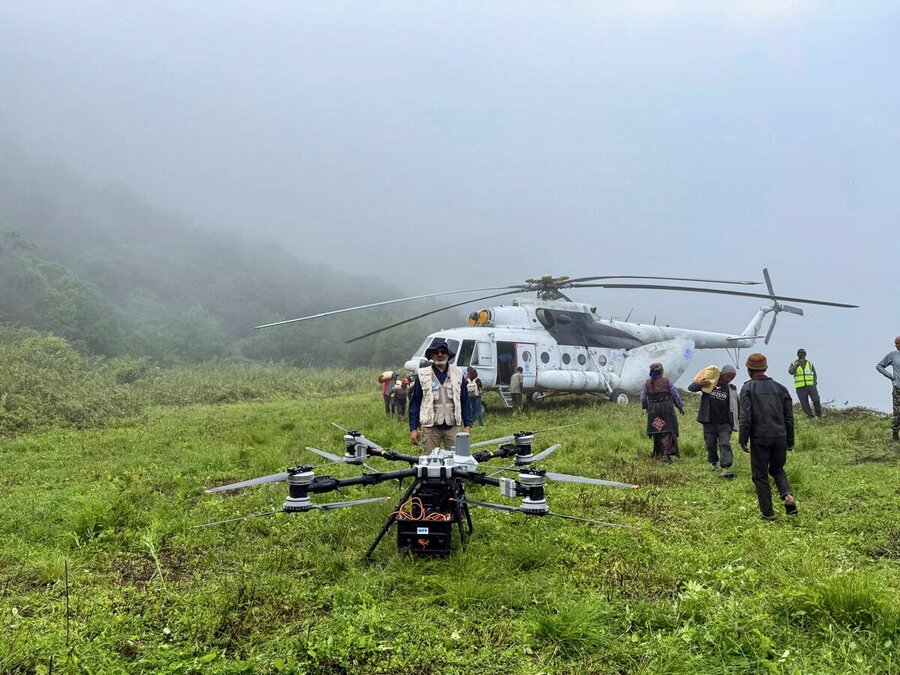 A man with a tan WFP vest and floppy hat stands on front of a humanitarian cargo drone, backdropped by green, fog-covered mountains, people and a helicopter. Photo: WFP/Photolibrary