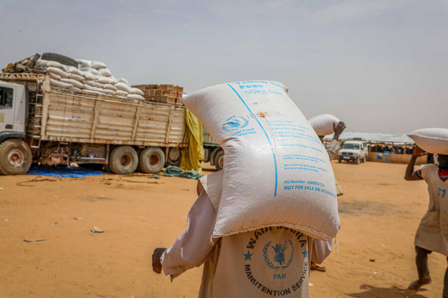 Workers transport bags of WFP food on their backs across a sandy landscape in Adre, Chad, backdropped by a WFP food truck. Photo: WFP/Arete/Salamon Djek