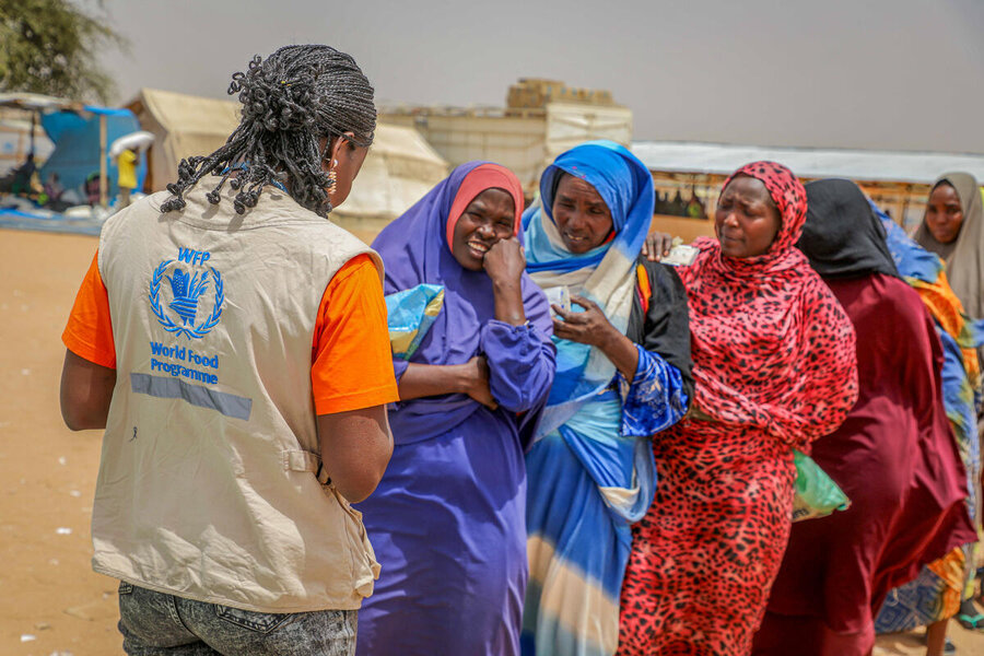 Women in colorful gowns and headscarves stand in a line as another woman in a tan WFP vest checks a list. Photo: WFP/Arete/Salamon Djek