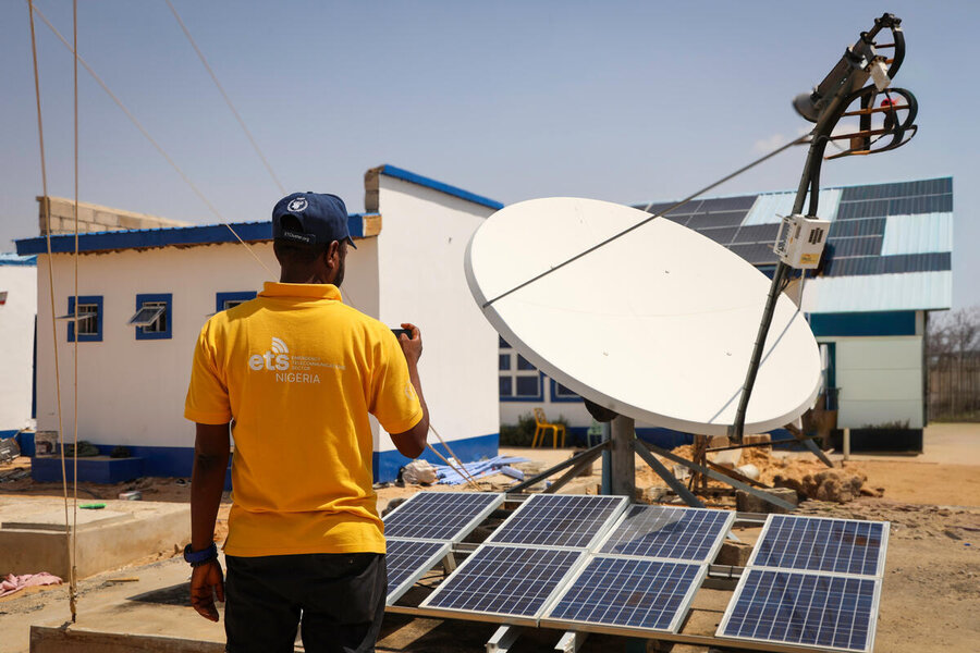A technician stands beside a satellite dish and solar panels at a communications site, inspecting connectivity equipment.