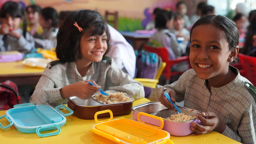 Two schoolchildren eat meals from lunchboxes while seated at a classroom table, with other students visible in the background during mealtime.