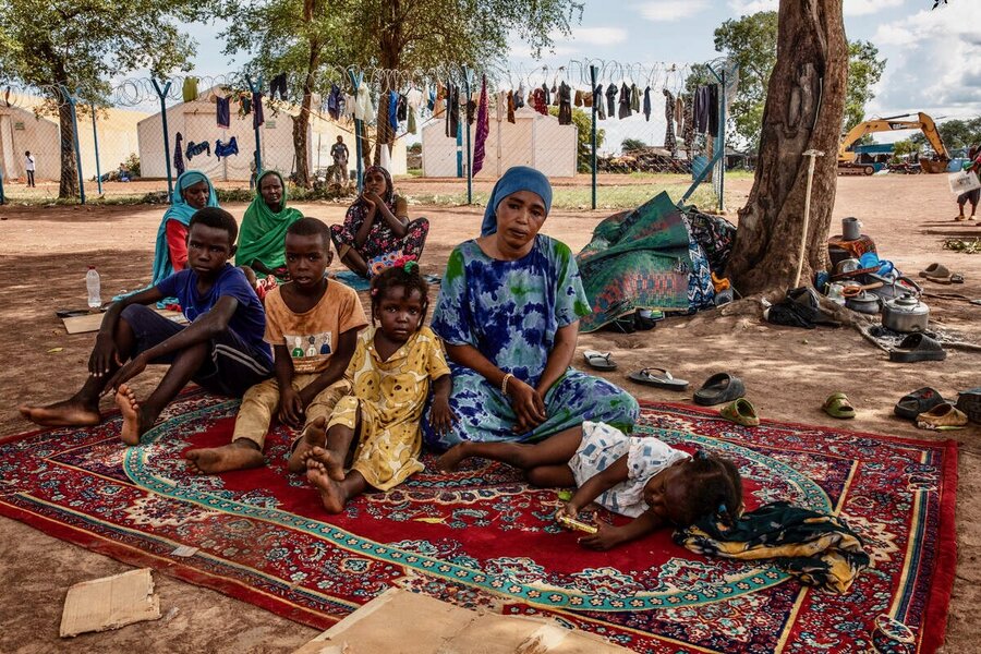 A woman in a colorful headscarf and dress sits on a carpet in a sandy refugee settlement with her children. Photo: WFP/Gabriela Vivacqua