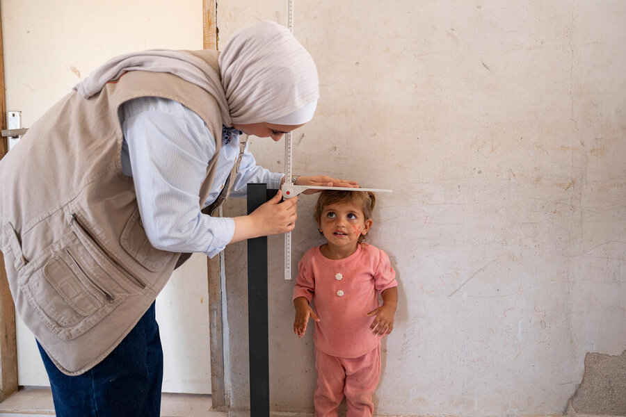 A woman in a headscarf and tan vest checks the size of a little girl in pink standing next to a wall. Photo: WFP/Hugh Rutherford