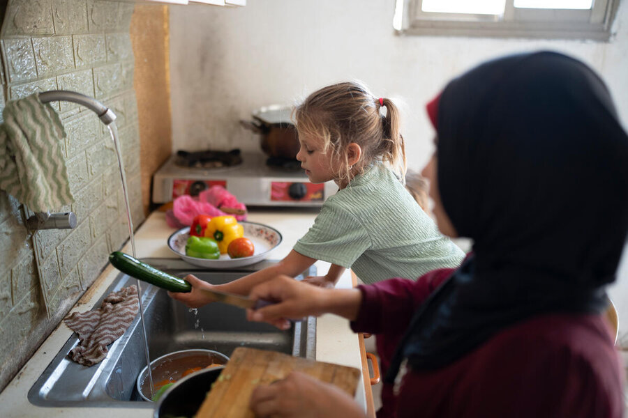 The back of a woman in a headscarf with a little girl next to her standing over a kitchen sink, washing vegetables. Photo: WFP/Hugh Rutherford